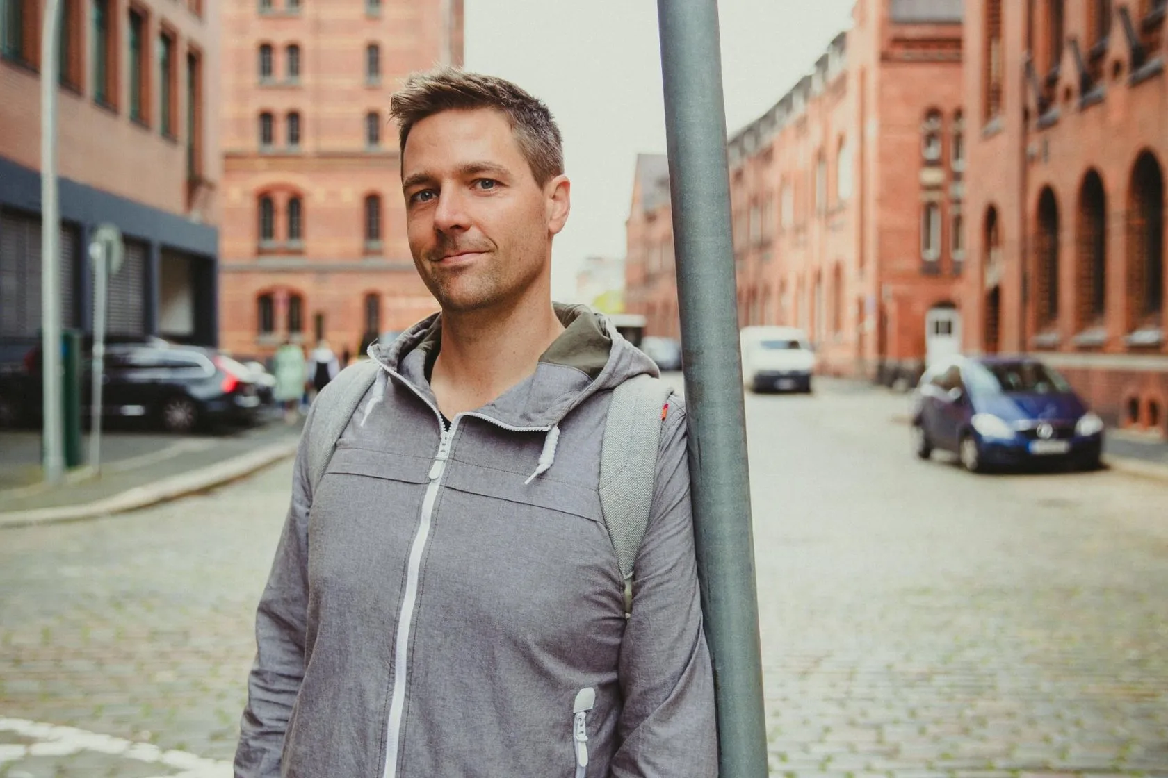 Frederic Hanusch leaning against a pole on a cobbled street between red-brick buildings
